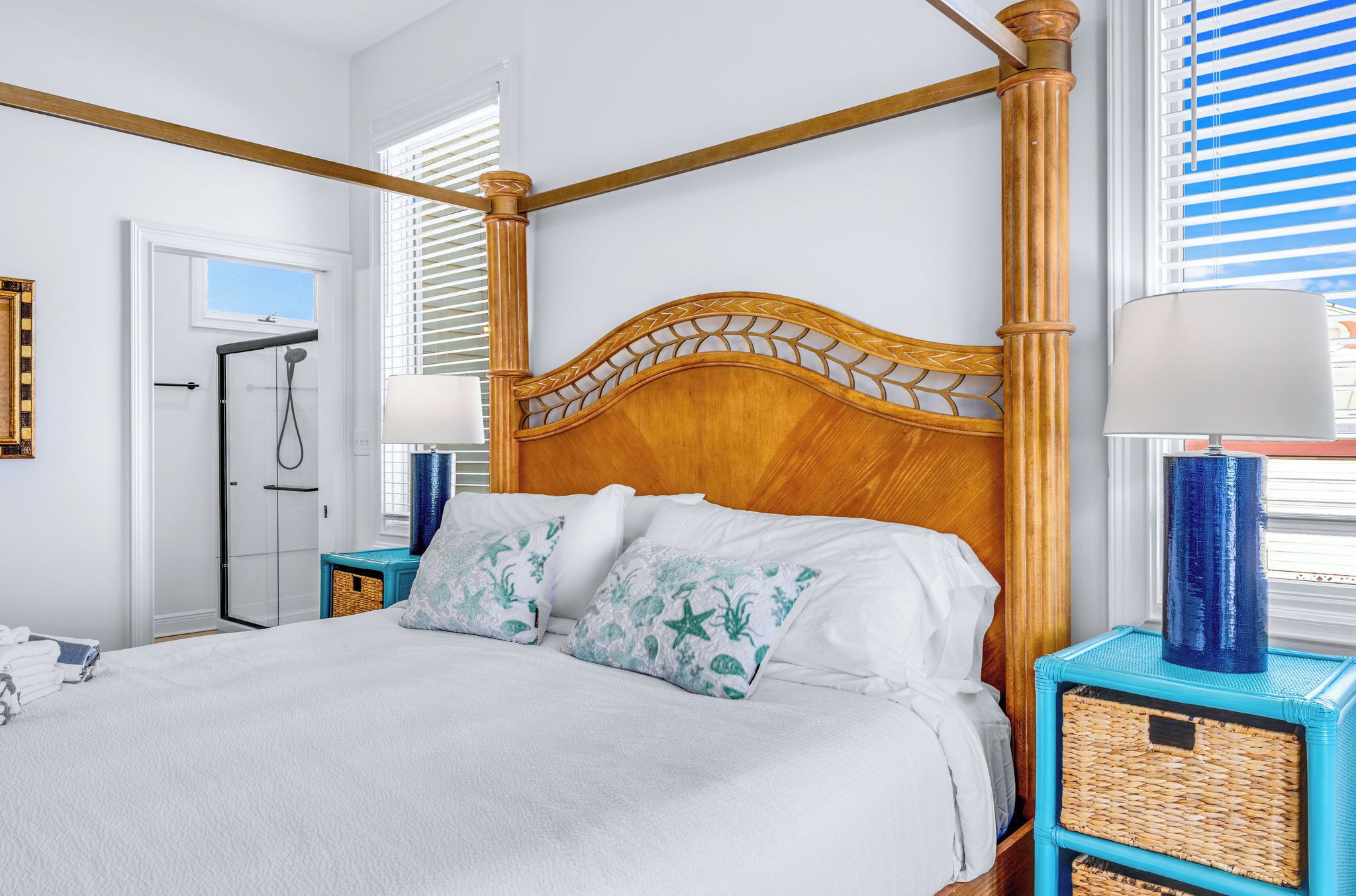 A bed with a wooden headboard, white bedding, and decorative pillows. There are two nightstands with lamps and wicker baskets. A walk-in closet and a door leading to a bathroom are visible.