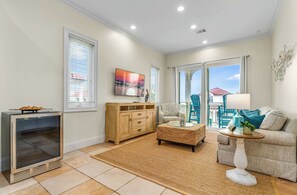 A living room with a television, a wooden cabinet, a wicker ottoman, a white table, and a sofa. - Salty Sunsets - Across from beach in exclusive Sunset Beach, coastal décor, views of Gulf and Bay! (St. George Island)