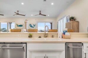 A modern kitchen with a stainless steel dishwasher, a double sink, and ceiling fans. - Salty Sunsets - Across from beach in exclusive Sunset Beach, coastal décor, views of Gulf and Bay! (St. George Island)