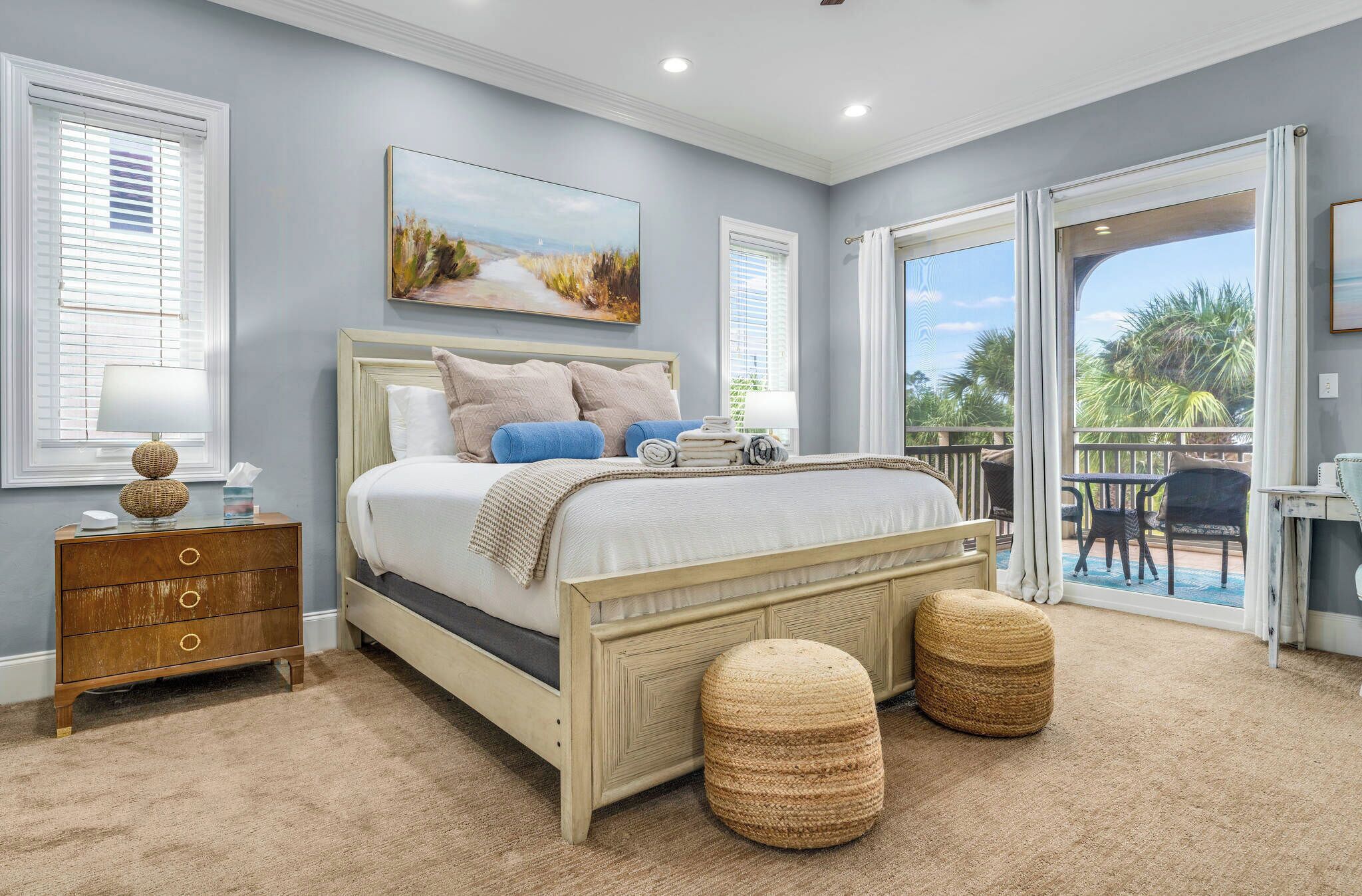 Une chambre à coucher comprenant un lit, des tables de chevet, une grande fenêtre et un balcon aménagé avec du mobilier d’extérieur.