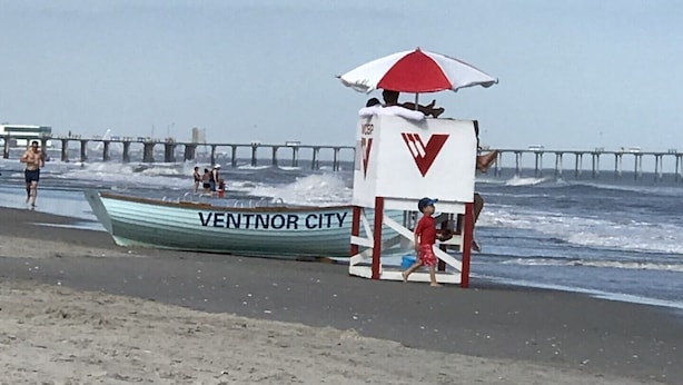 Ein Strand mit einem Rettungsturm, einem Steg im Hintergrund und einem Boot mit der Aufschrift 'VENTNOR CITY' im Sand.