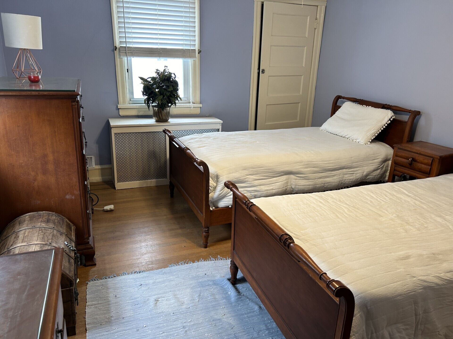 Un dormitorio con dos camas de madera, un aparador, una mesita de noche, una ventana con persianas y un radiador.