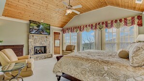 Ein Schlafzimmer mit Kamin aus Stein, einem Bett mit gemusterten Textilien, einem Glastisch, einer Holzbalkendecke und einem an der Wand montierten Fernseher.