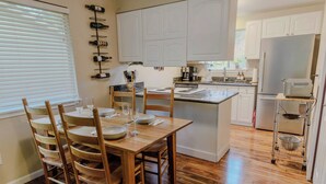 Una cocina con suelo de madera, armarios blancos, una mesa de comedor con sillas y una estantería para vinos en la pared.