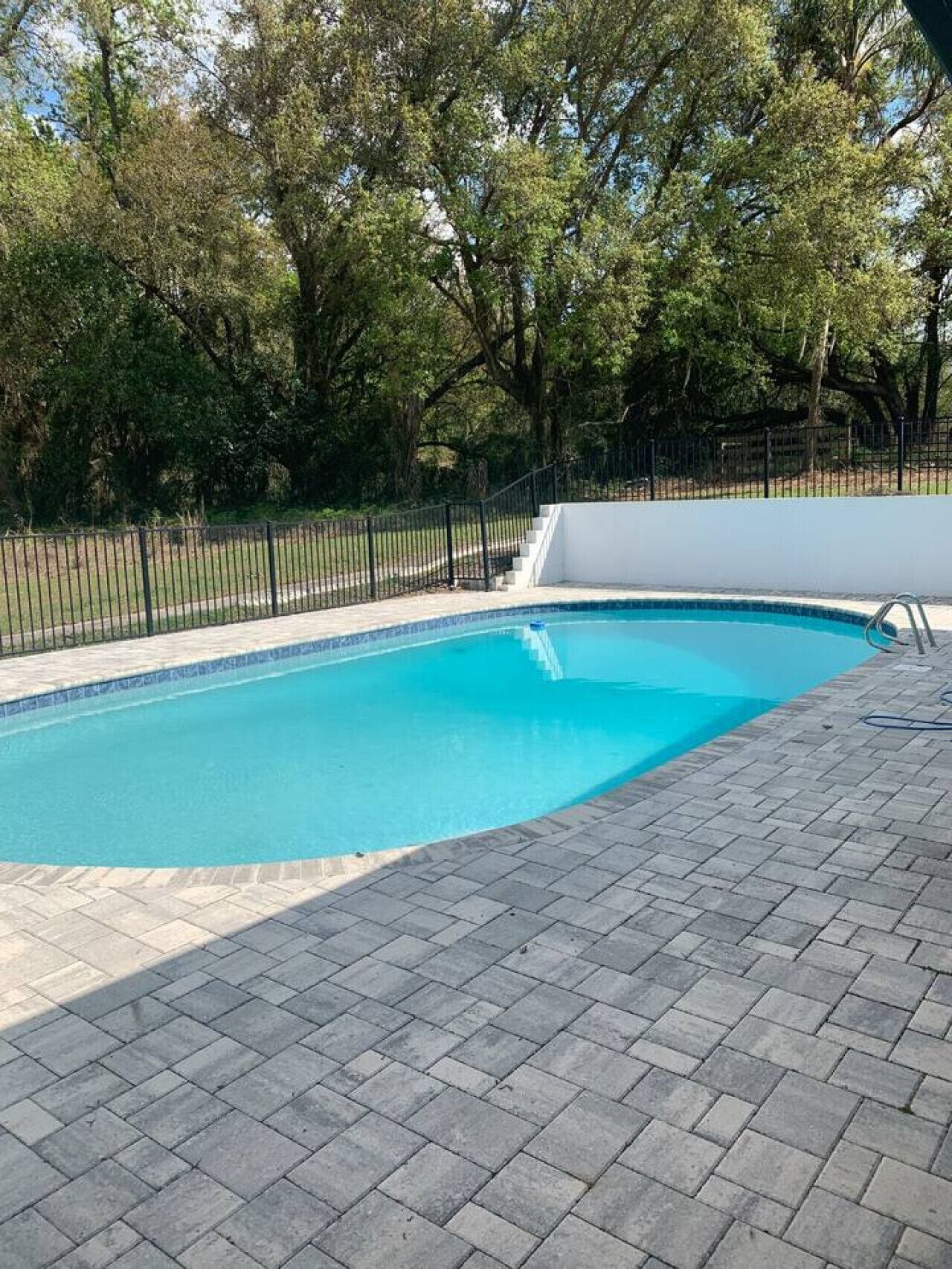 A swimming pool with a tiled deck, surrounded by a fence and trees.