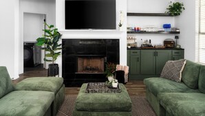 A living room with a fireplace, green sofas, a flat-screen TV, and a kitchen area with shelves. - Geyser Ave (Pflugerville)
