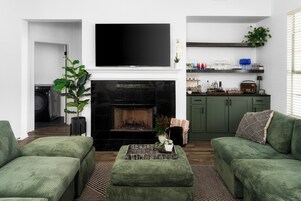Una sala de estar con chimenea, sofás verdes, un televisor de pantalla plana y una zona de cocina con estantes.