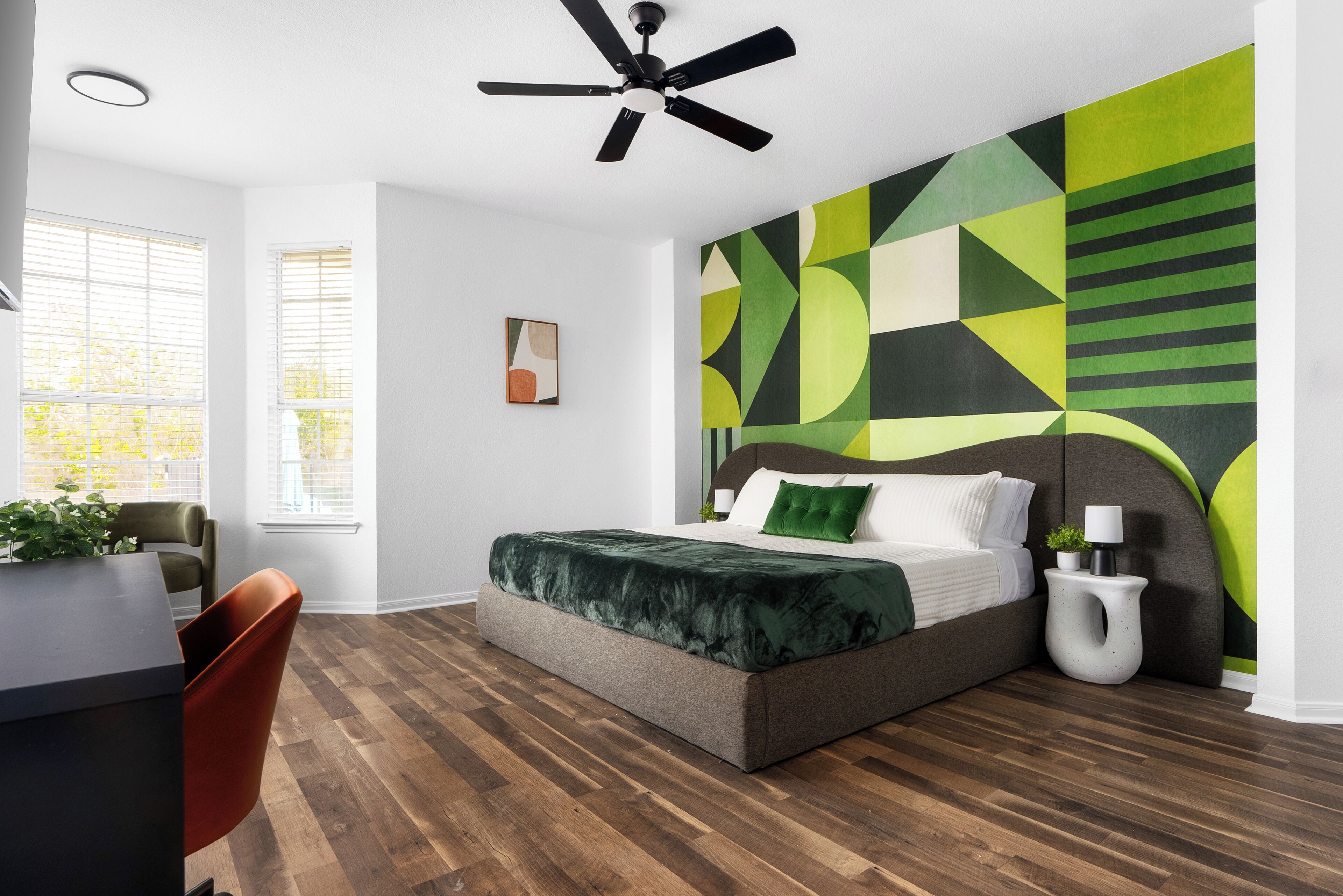 Een slaapkamer met een bed, een plafondventilator, een muur met geometrische patronen, een raam met zonwering en een plant.