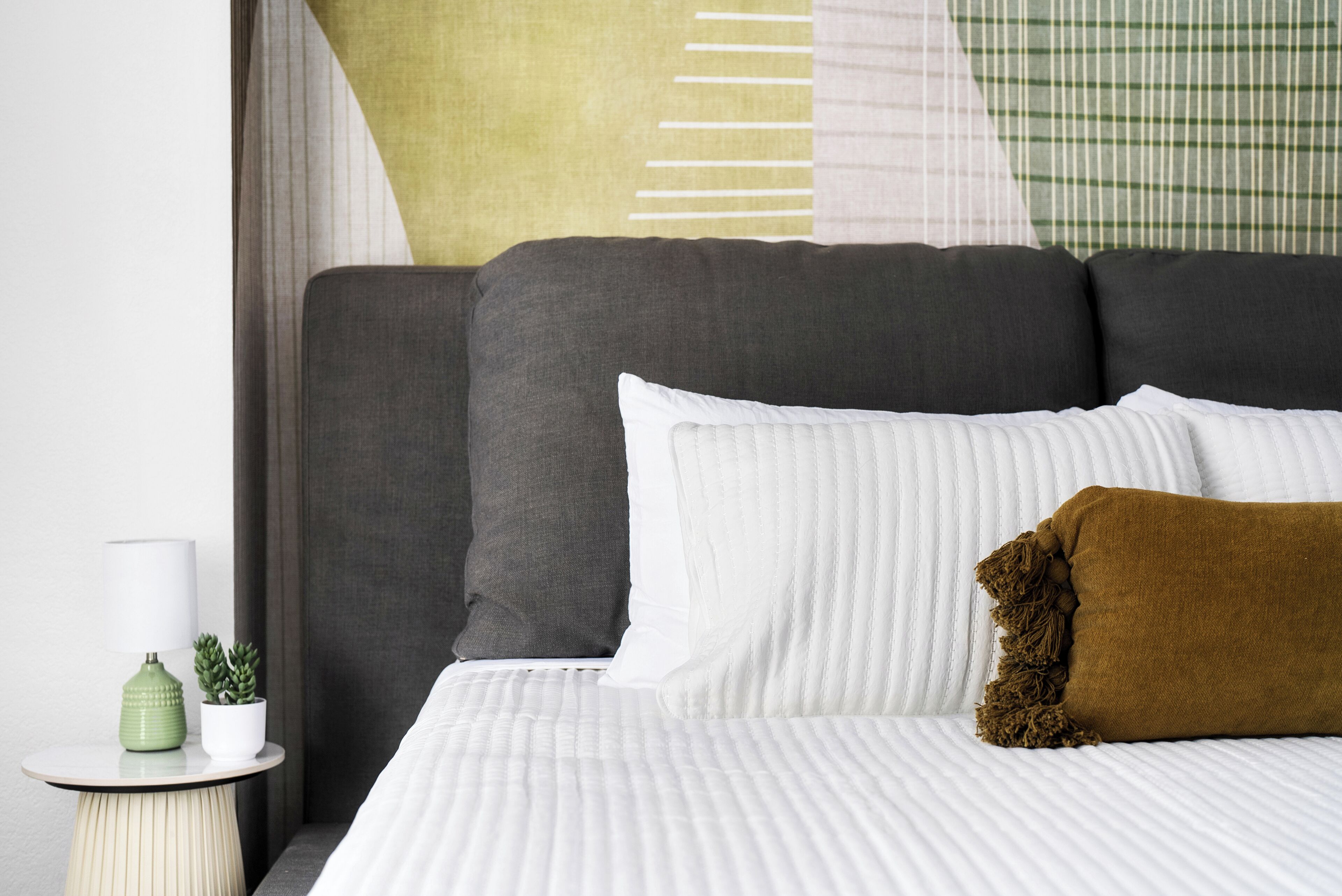 Un letto con biancheria da letto bianca, un cuscino marrone e un comodino con una lampada verde e una pianta in vaso.