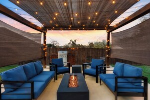 Un patio al aire libre con una chimenea, asientos acolchados azules y luces colgantes.