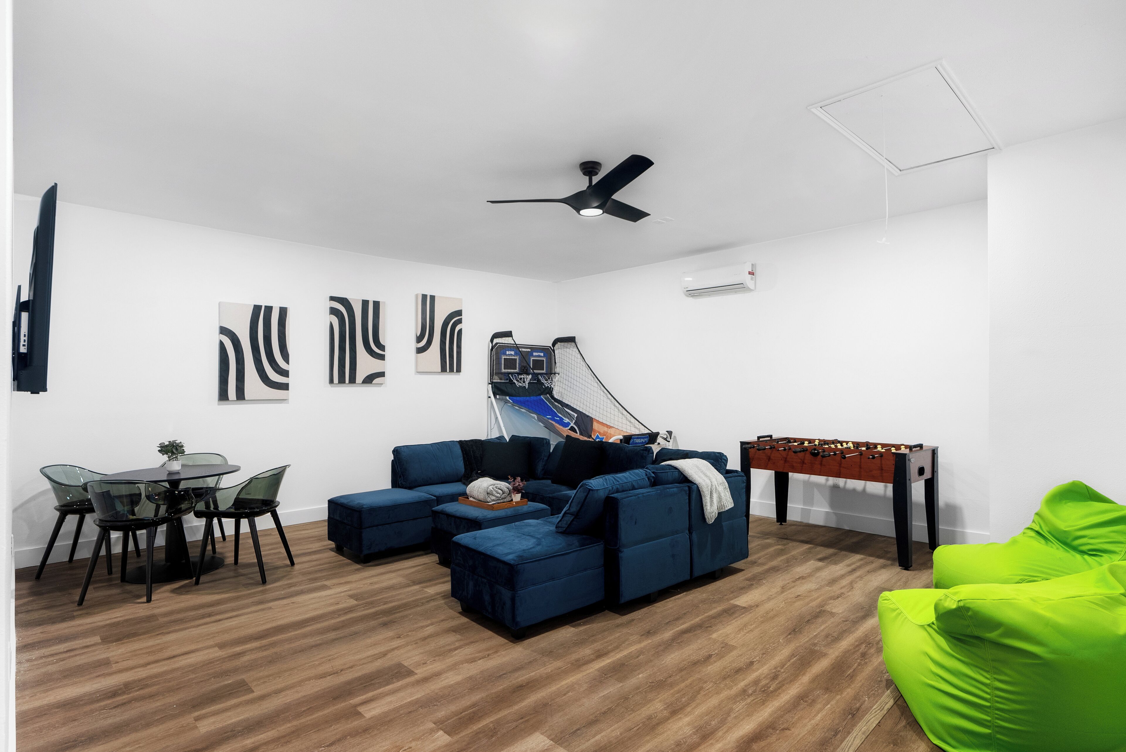 A living room with a blue sofa, a bright green chair, a foosball table, a ceiling fan, and abstract wall art.