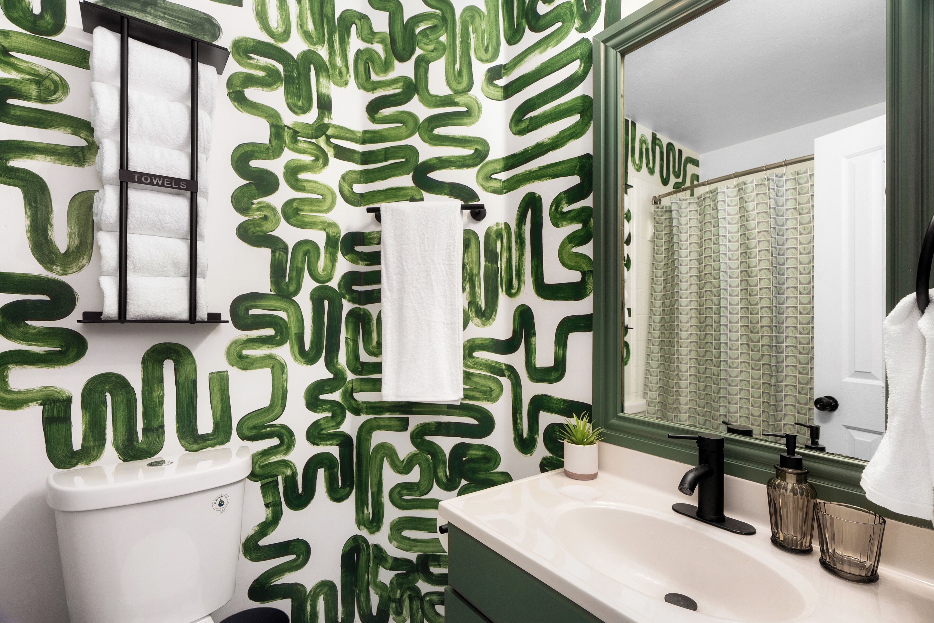 A bathroom with a green abstract wall design, a white sink, a black faucet, and a glass shower curtain.