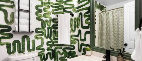 A bathroom with a green abstract wall design, a white sink, a black faucet, and a glass shower curtain.