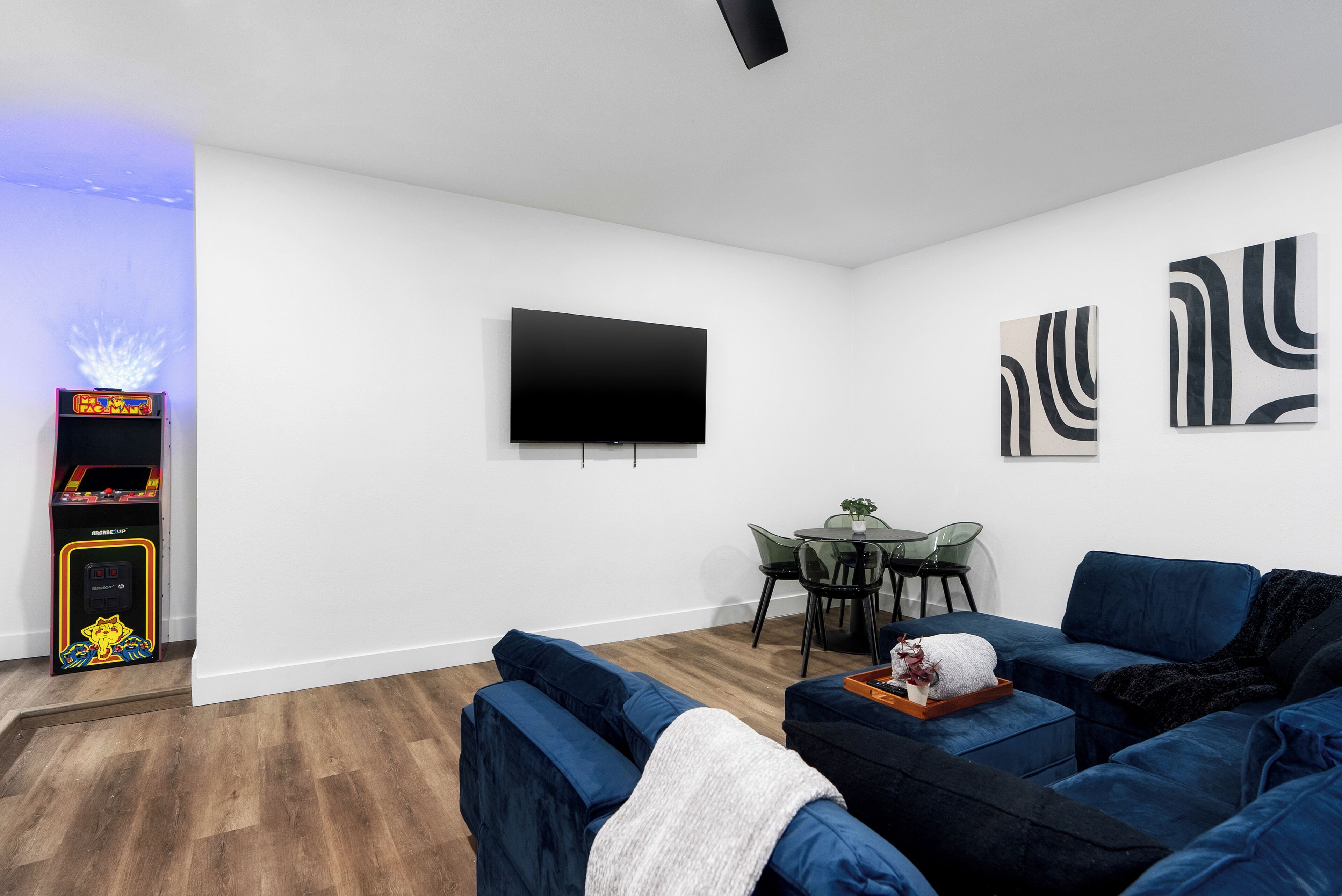 A living room with a flat-screen TV, blue sofas, a small table with chairs, and a gaming machine.