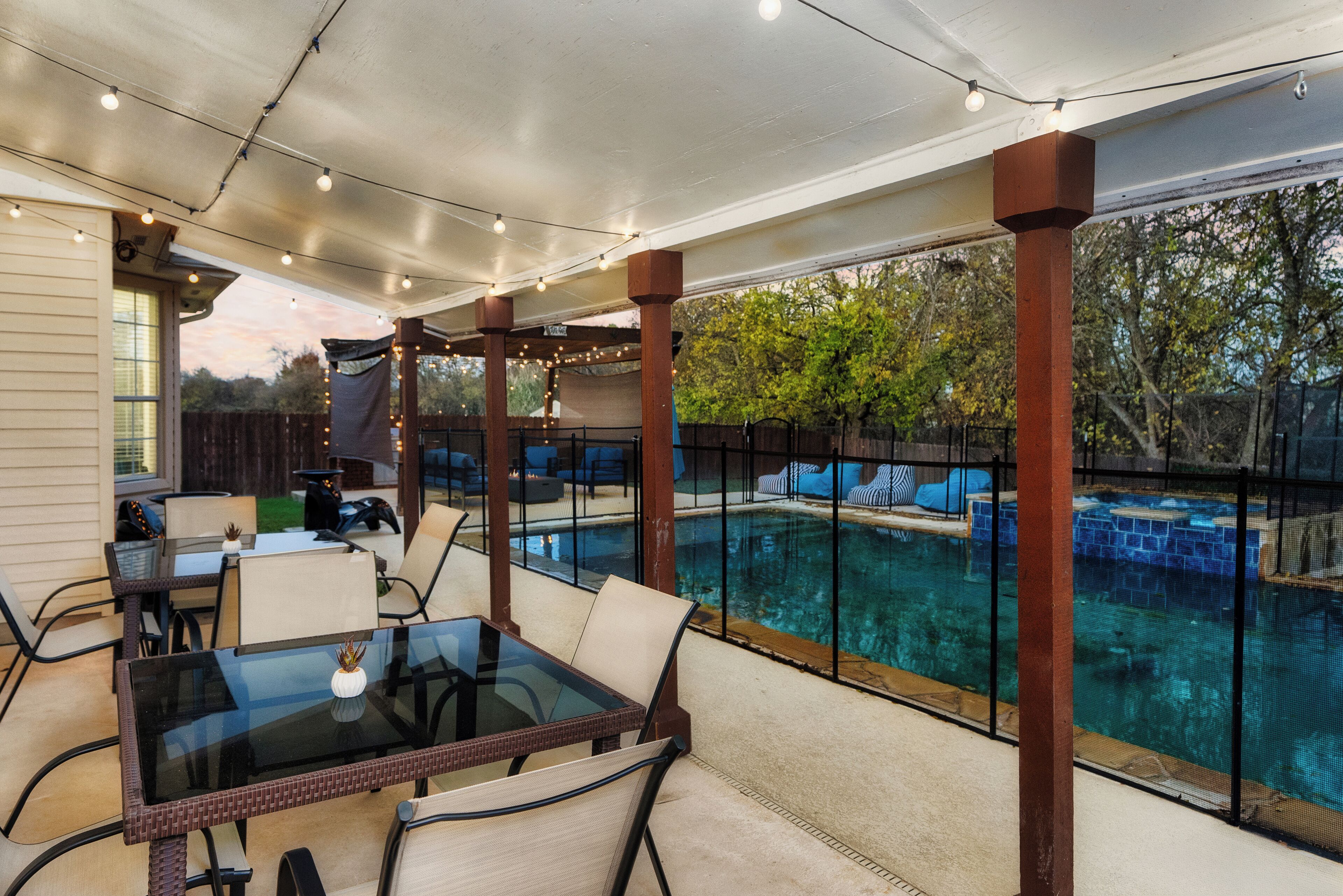 A covered patio with a glass table and chairs, a pool, and a view of trees.