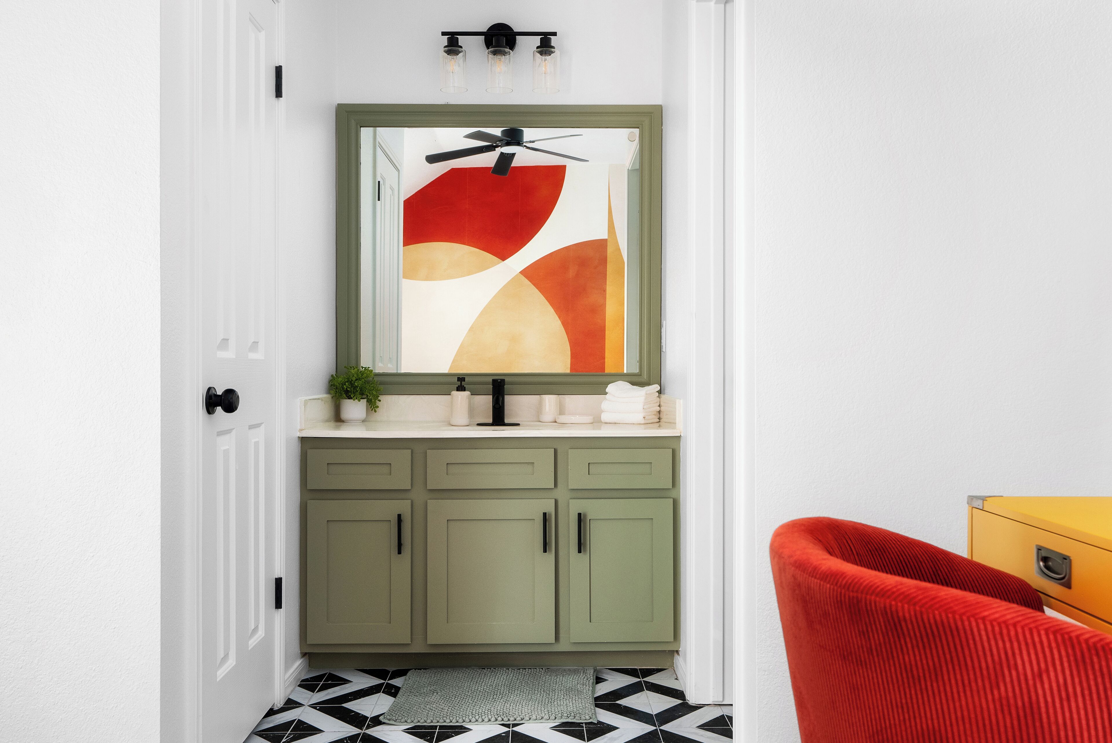 A bathroom with a green vanity, a ceiling fan, a large abstract painting, and a red chair.