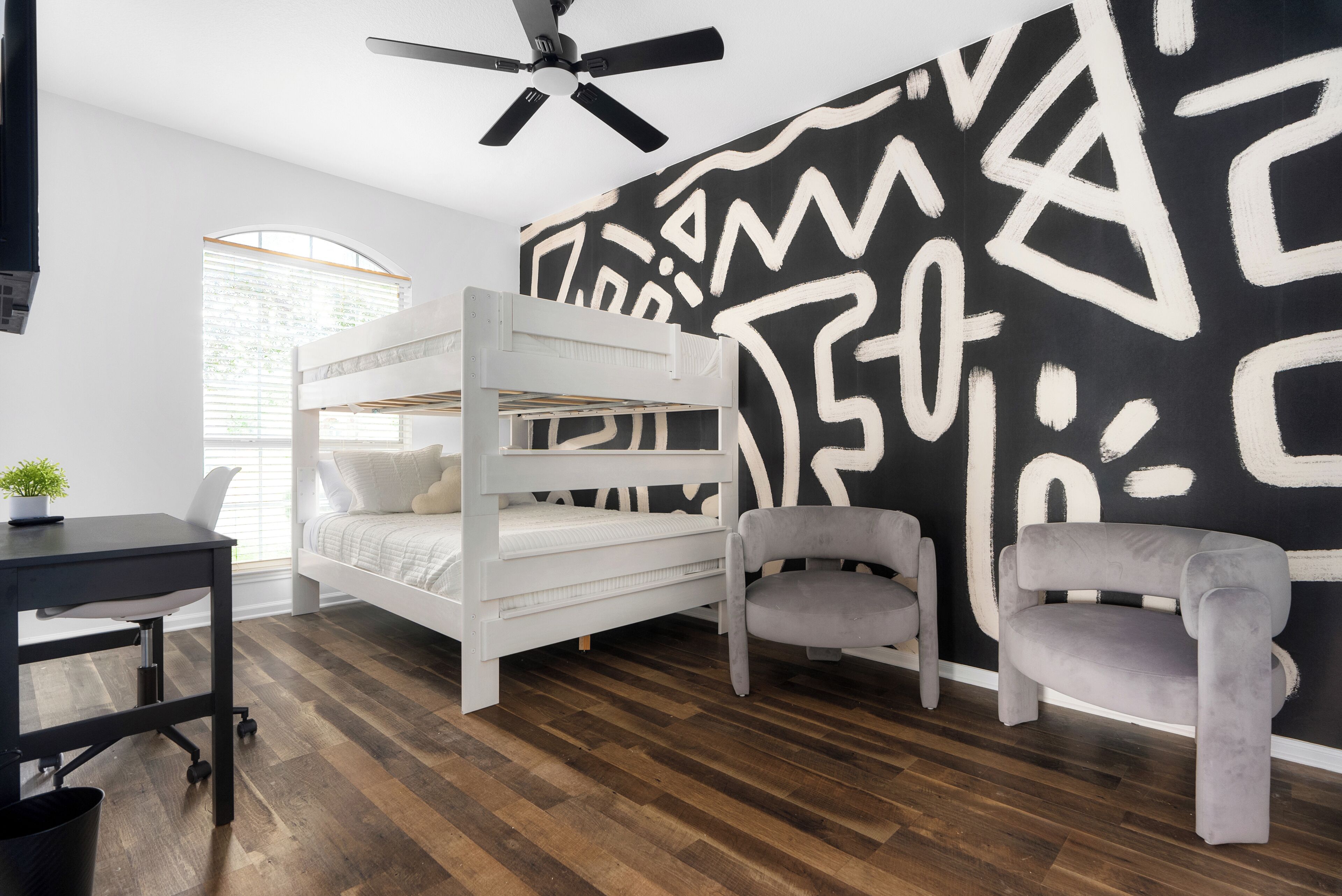 A room with a bunk bed, a desk, a chair, a ceiling fan, and a black and white abstract wall.