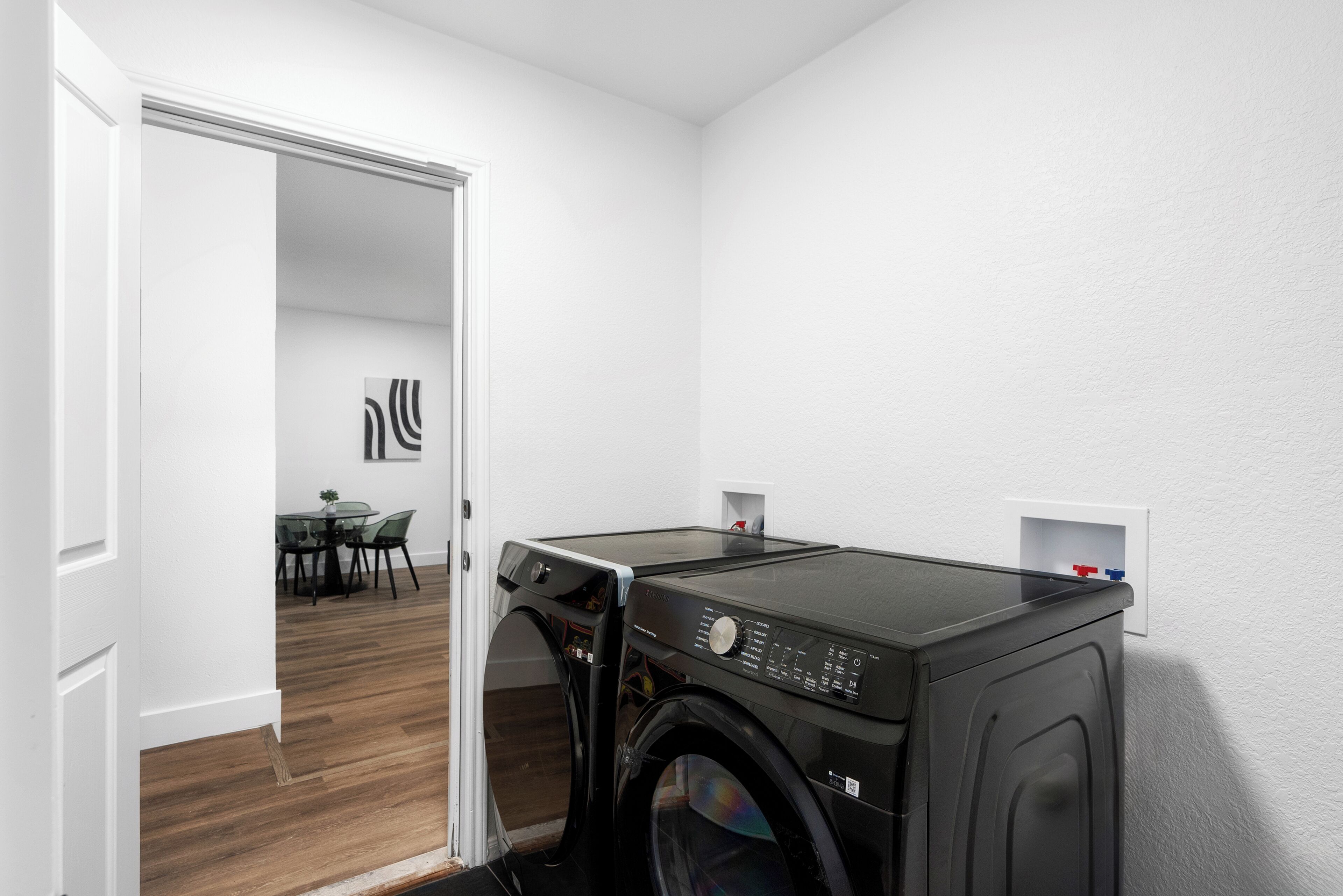 A laundry room with a washing machine and dryer, a dining area with chairs and a table, and a white wall with a small shelf.