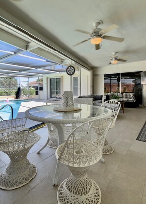 Eine überdachte Terrasse mit einem Glastisch und weißen Korbstühlen, einem Deckenventilator und einem Pool im Hintergrund.