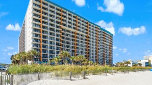 A high-rise hotel with multiple balconies, located near a beach with a wooden fence and palm trees. - Landmark Resort Ocean View | Beach, Pools, Lazy River (Myrtle Beach)