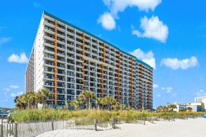 A high-rise hotel with multiple balconies, located near a beach with a wooden fence and palm trees. - Landmark Resort Ocean View | Beach, Pools, Lazy River (Myrtle Beach)