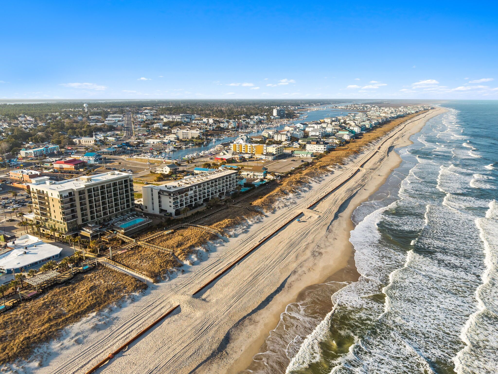 Vue aérienne d’une ville côtière avec un hôtel en bord de mer, une marina et une vaste plage de sable.