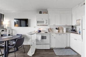 A modern kitchen with white cabinetry, a microwave, an oven, and a dining area with a table and chairs. - Boardwalk Breeze, Oceanfront, Pool! Villa (Carolina Beach)