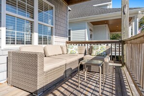 Uma área de estar coberta ao ar livre, com um sofá de vime e uma mesa pequena, ao lado de uma casa com janelas amplas e um deck de madeira.
