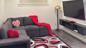 Un salon moderne avec un canapé gris, des coussins rouges en forme de cœur, un tapis à motifs, un téléviseur à écran plat et une lampe sur pied.