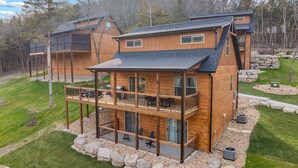 A two-story wooden cabin with a balcony, stone foundation, and a clear view of the surrounding forest. - New Cozy Lodge w Arcade_Private Hot Tub_Cmty In/Outdoor Pools & Much More_Free Attraction Tickets (Hollister)