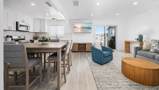 A modern kitchen with white cabinets, a dining area with a wooden table and chairs, a living room with a blue sofa and a wooden coffee table, and a view of the outside through a glass door. - Balboa Remodeled Upstairs Unit with Parking Near the Funzone (Newport Beach)