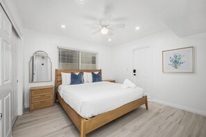 Une chambre à coucher comprenant un lit, un ventilateur de plafond, une table de chevet, un miroir, une fenêtre avec des stores, une porte et un tableau encadré accroché au mur.