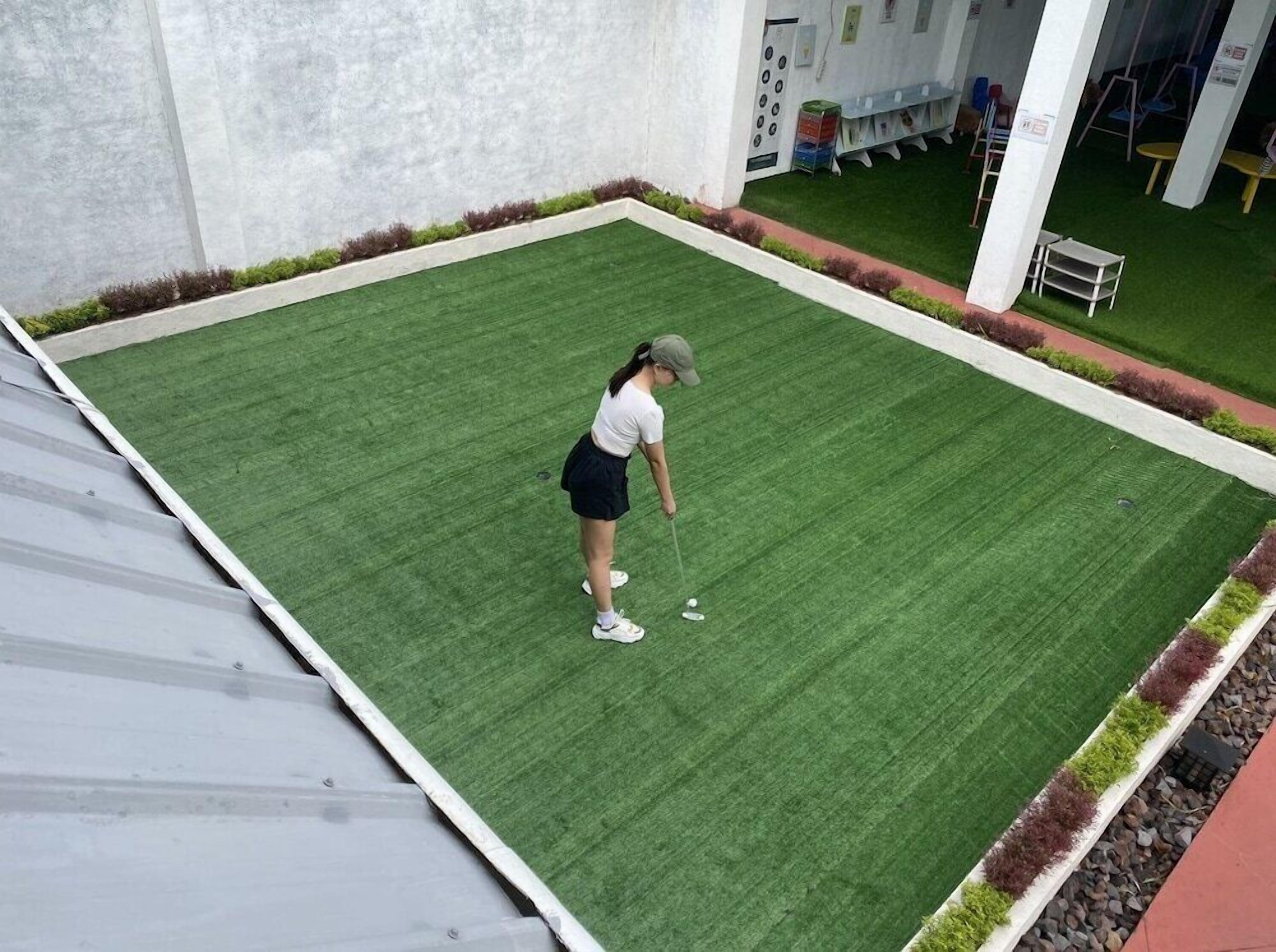 Una persona juega al golf sobre un campo verde, con un muro blanco y un edificio al fondo.