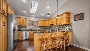 A kitchen with wooden cabinets, a stainless steel refrigerator, a stone backsplash, and a window with a view of trees. - Scenic Solitude in Lake Lure (Lake Lure)