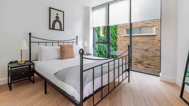 Quarto moderno com estrutura de cama em metal, mesa de cabeceira com abajur, janela grande com vista para vegetação e parede de tijolos.