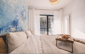 Ein Schlafzimmer mit einem Bett, einem Nachttisch mit einem Tablett Croissants, Blick ins Freie durch ein Fenster mit Vorhängen und einer strukturierten Wand.