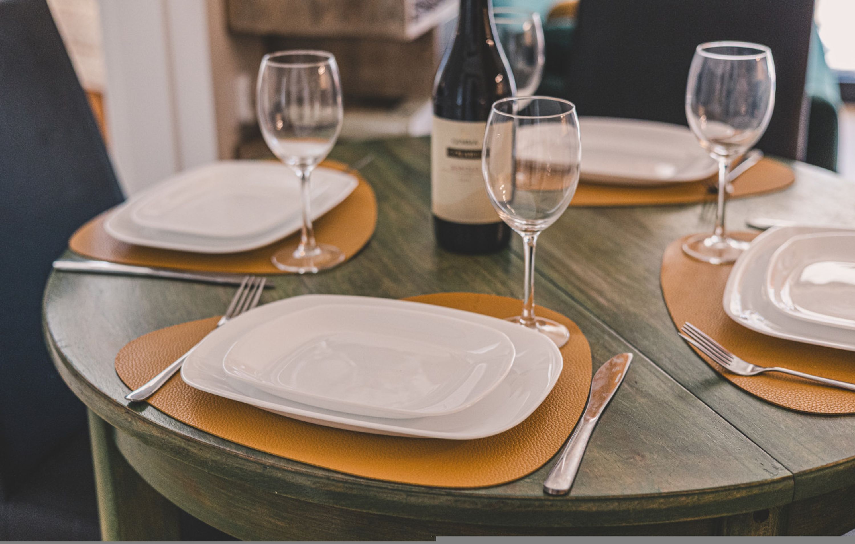 Una mesa de comedor dispuesta con platos, cubiertos y copas de vino, junto con una botella de vino y un mantel individual.