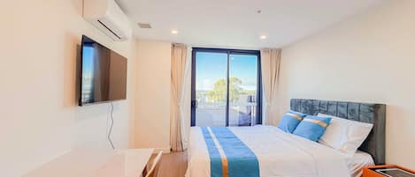 A hotel room with a bed, a TV, an air conditioning unit, a window with curtains, and a view of the outside.