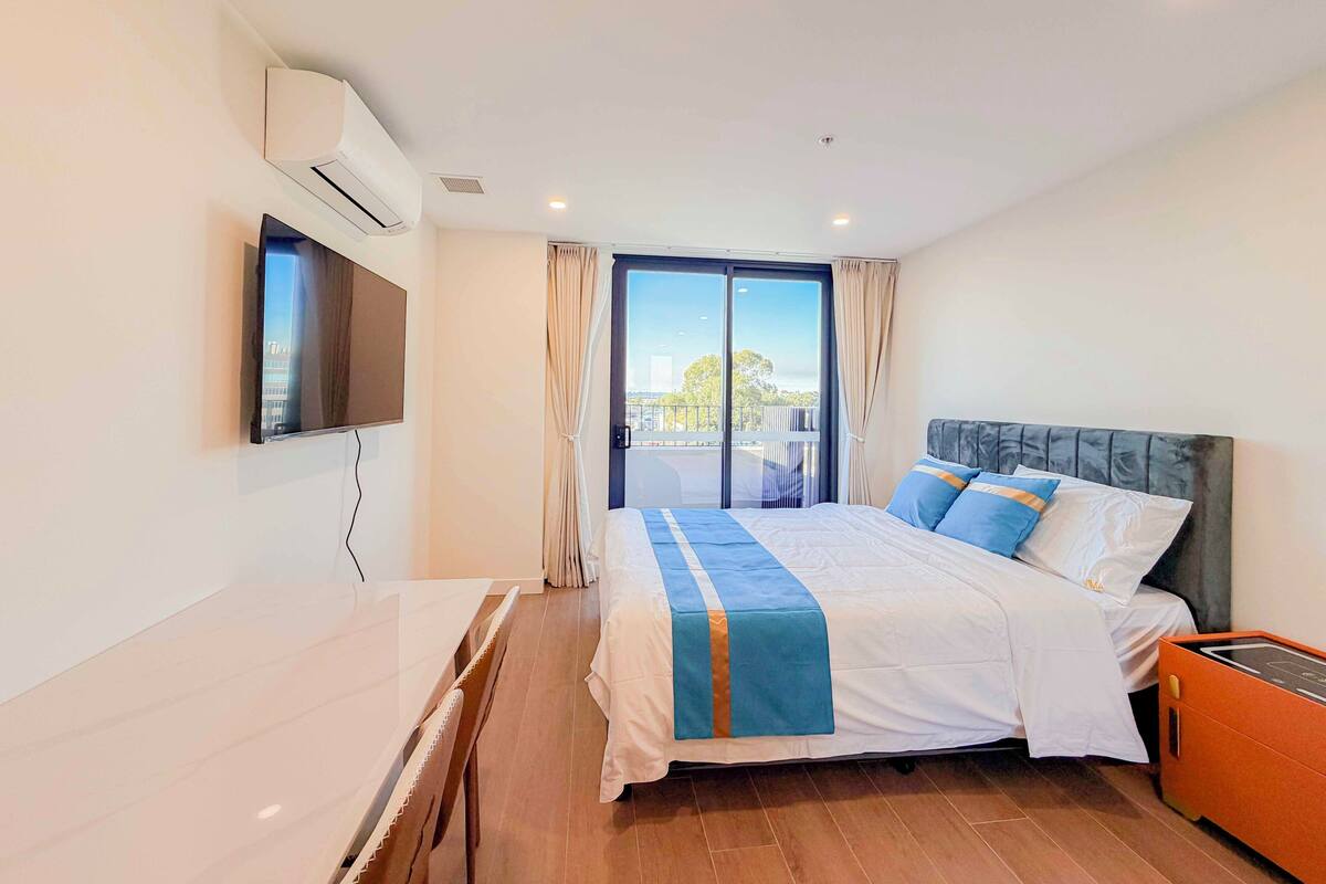 A hotel room with a bed, a TV, an air conditioning unit, a window with curtains, and a view of the outside.