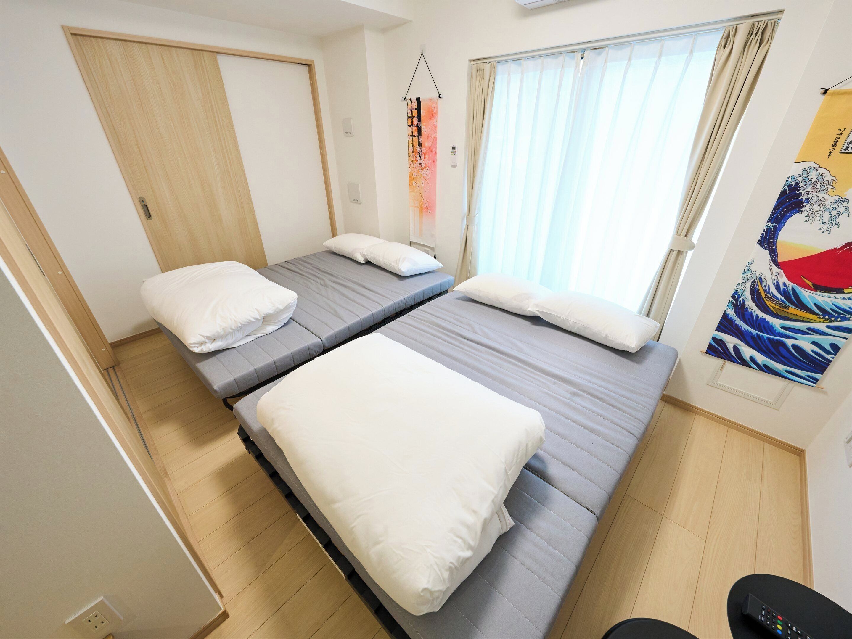 A room with two beds, a wooden floor, a window with curtains, and a wall with a Japanese artwork.