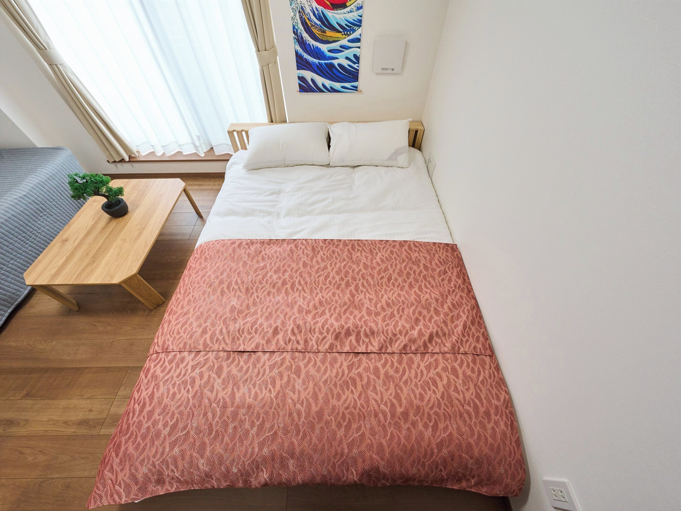 A single bed with a white and pink patterned bedspread, a small wooden table with a potted plant, and a wall-mounted air conditioner.