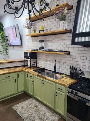 Una cocina con armarios verdes, una encimera de madera, un lavamanos de acero inoxidable, una estufa y un televisor montado en la pared.