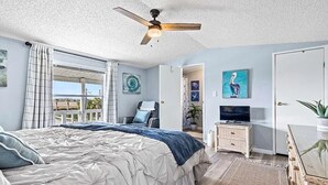 A bedroom with a bed, a ceiling fan, a television, a dresser, and a door leading to another room.