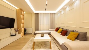 A modern living room with a flat-screen TV, a sofa, a coffee table, and a large window with curtains. - Villa Royale in Durr s (Durrës)