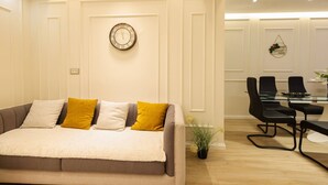 A modern room with a sofa, a dining table with chairs, a wall clock, and a small plant. - Villa Royale in Durr s (Durrës)