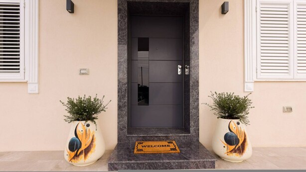 A dark door with a welcome mat, two potted plants with abstract designs, and a light-colored wall. - Villa Royale in Durr s (Durrës)