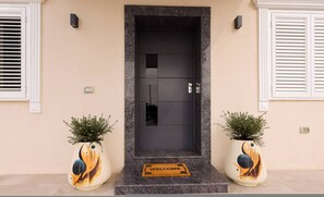 Une porte sombre avec un paillasson, deux plantes en pot ornées de motifs abstraits et un mur de couleur claire.