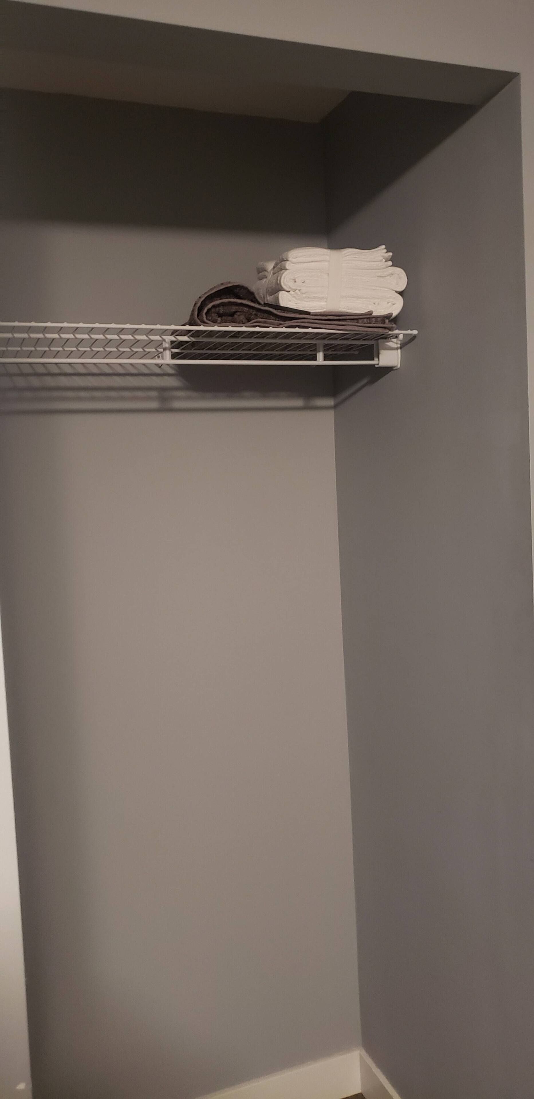 A corner of a room with a grey wall and a metal shelf holding folded towels.