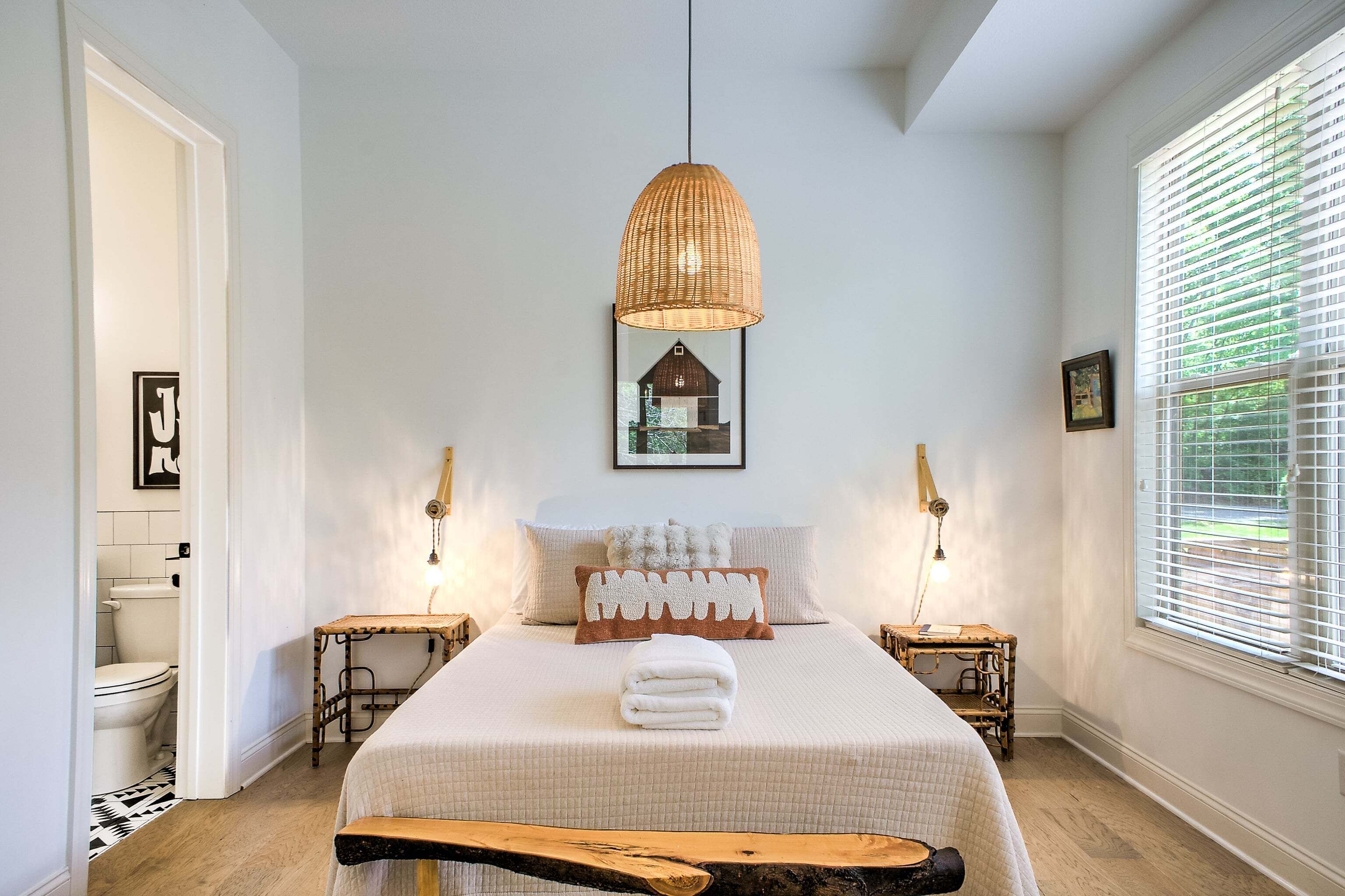 A bedroom with a bed, a hanging light fixture, a bedside table, a window with blinds, and a framed picture on the wall.