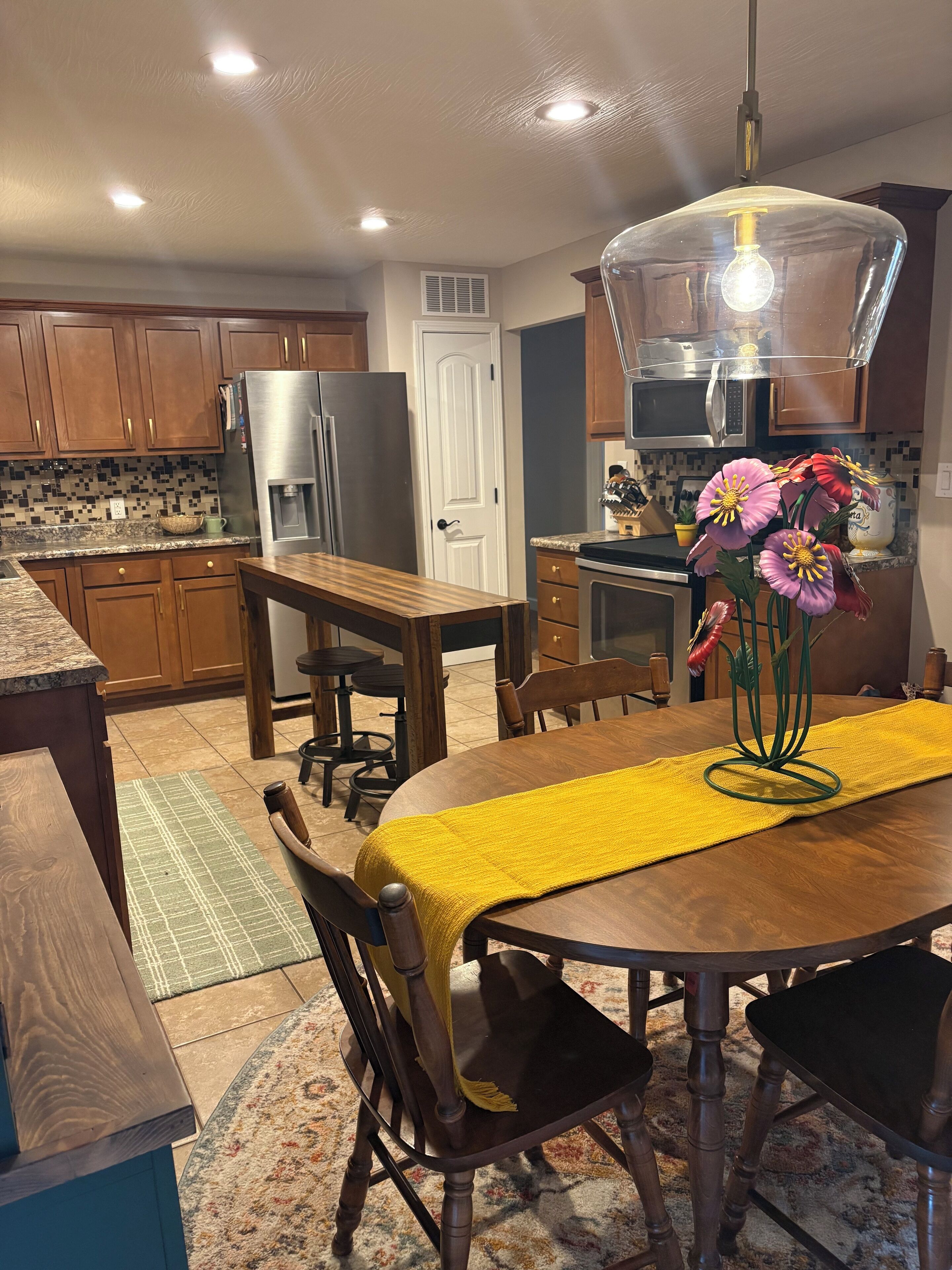 A kitchen with wooden cabinets, a stainless steel refrigerator, a dining table with a yellow table runner, and a flower vase.